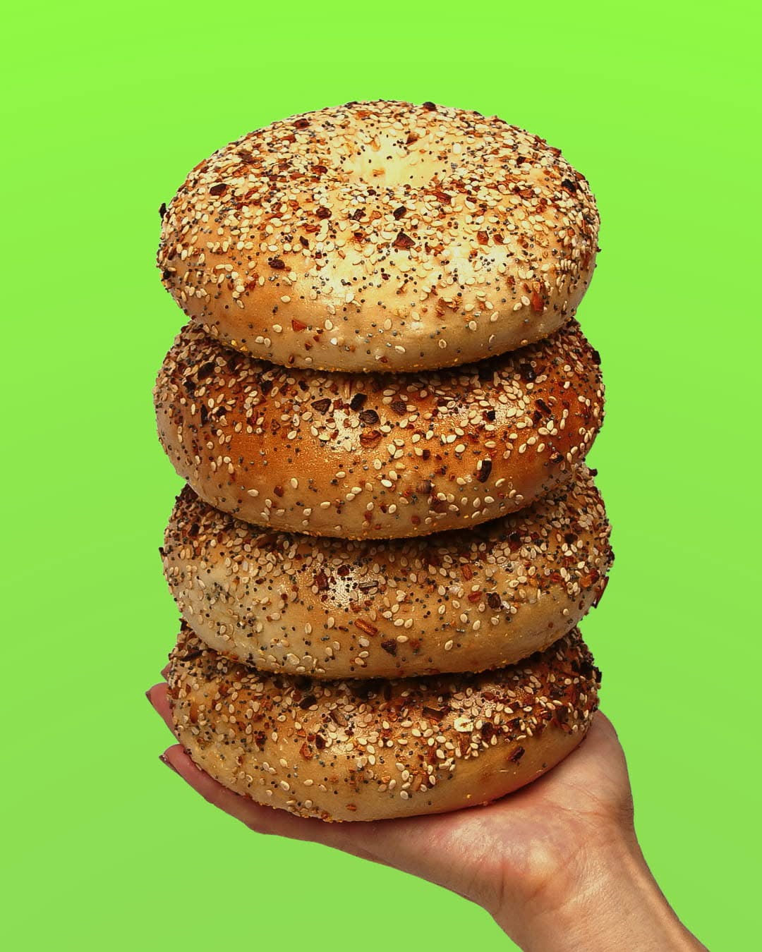 A hand holding a stack of four everything bagels on a bright green background